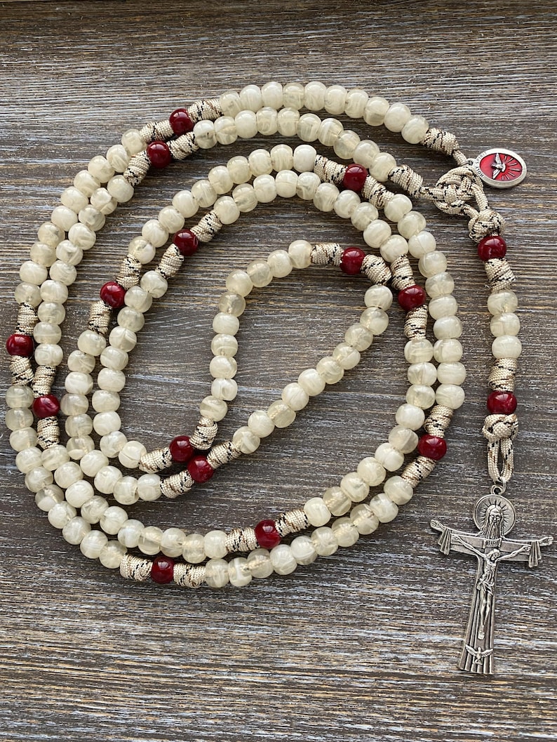 Puede incluir: Un rosario con cuentas transl&uacute;cidas de color blanco roto y cuentas m&aacute;s peque&ntilde;as de color rojo oscuro ensartadas en un cord&oacute;n trenzado. La pieza incluye un crucifijo plateado con la figura de Jes&uacute;s y una peque&ntilde;a medalla ovalada con una paloma roja.