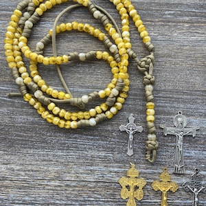 Puede incluir: Un rosario hecho de cuentas transl&uacute;cidas amarillas y blancas ensartadas en un cord&oacute;n verde oliva, con un dise&ntilde;o anudado. Junto al rosario hay cinco colgantes de crucifijo de metal en tonos plateado, dorado y gris oscuro, cada uno con una figura en la cruz.