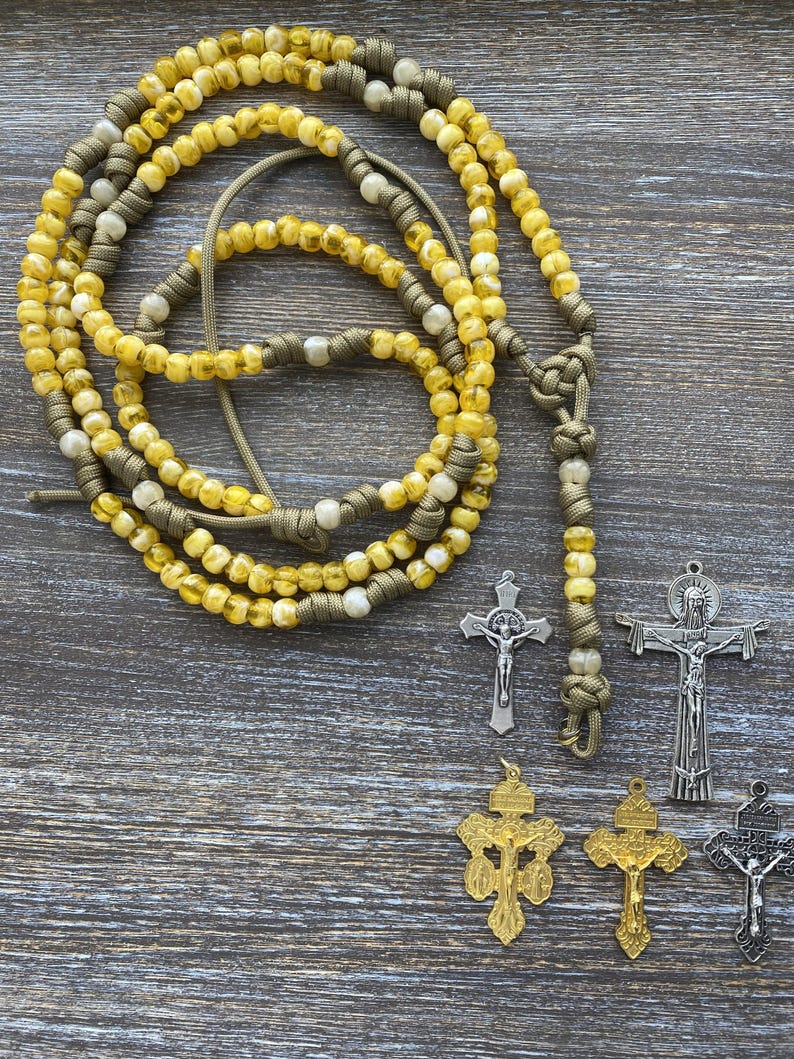Puede incluir: Un rosario hecho de cuentas transl&uacute;cidas amarillas y blancas ensartadas en un cord&oacute;n verde oliva, con un dise&ntilde;o anudado. Junto al rosario hay cinco colgantes de crucifijo de metal en tonos plateado, dorado y gris oscuro, cada uno con una figura en la cruz.