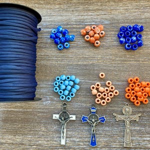 Puede incluir: Una bobina grande de cord&oacute;n de nailon azul oscuro acompa&ntilde;ada de varias cuentas sueltas en tonos azul, naranja y melocot&oacute;n, junto con tres colgantes de crucifijo de metal, incluido uno con detalles en esmalte azul y otro con la inscripci&oacute;n INRI.