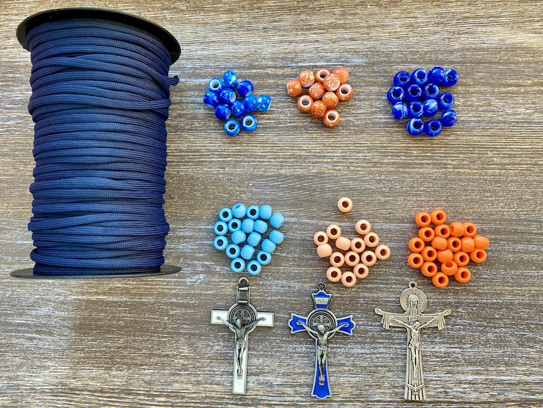 Puede incluir: Una bobina grande de cord&oacute;n de nailon azul oscuro acompa&ntilde;ada de varias cuentas sueltas en tonos azul, naranja y melocot&oacute;n, junto con tres colgantes de crucifijo de metal, incluido uno con detalles en esmalte azul y otro con la inscripci&oacute;n INRI.