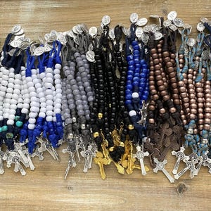 Puede incluir: Una colecci&oacute;n de m&uacute;ltiples rosarios con cuentas de varios colores, incluyendo blanco, azul, negro, gris y bronce met&aacute;lico. Cada rosario est&aacute; construido con cord&oacute;n anudado, un crucifijo de metal y una peque&ntilde;a medalla religiosa circular.