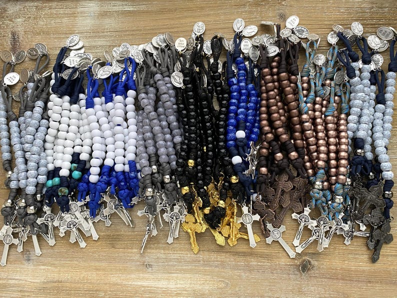 Puede incluir: Una colecci&oacute;n de m&uacute;ltiples rosarios con cuentas de varios colores, incluyendo blanco, azul, negro, gris y bronce met&aacute;lico. Cada rosario est&aacute; construido con cord&oacute;n anudado, un crucifijo de metal y una peque&ntilde;a medalla religiosa circular.
