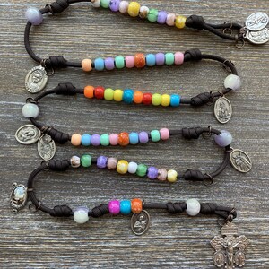 Puede incluir: Un rosario hecho de paracord marr&oacute;n oscuro con secciones de cuentas de pl&aacute;stico multicolores en tonos rosa, azul, verde, amarillo y naranja. El rosario incluye varias medallas religiosas de metal plateado y un crucifijo en el extremo.