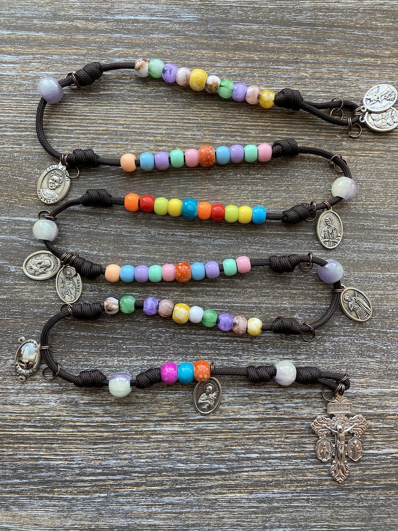 Puede incluir: Un rosario hecho de paracord marr&oacute;n oscuro con secciones de cuentas de pl&aacute;stico multicolores en tonos rosa, azul, verde, amarillo y naranja. El rosario incluye varias medallas religiosas de metal plateado y un crucifijo en el extremo.
