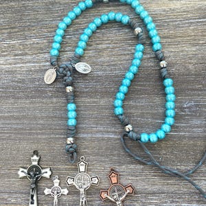 Puede incluir: Un rosario con cuentas de color turquesa brillante ensartadas en un cord&oacute;n gris con detalles de metal plateado y dos peque&ntilde;as medallas ovaladas. Debajo del rosario hay cuatro crucifijos distintos con diversos acabados met&aacute;licos, incluyendo plata, negro y cobre.