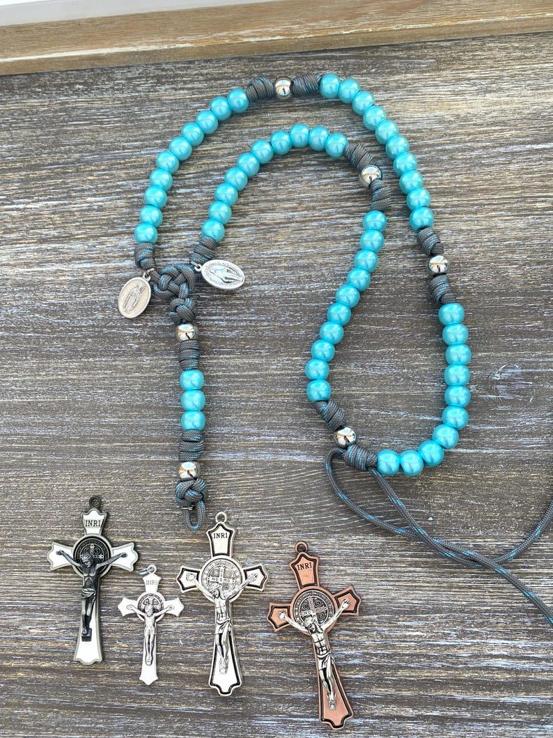 Puede incluir: Un rosario con cuentas de color turquesa brillante ensartadas en un cord&oacute;n gris con detalles de metal plateado y dos peque&ntilde;as medallas ovaladas. Debajo del rosario hay cuatro crucifijos distintos con diversos acabados met&aacute;licos, incluyendo plata, negro y cobre.