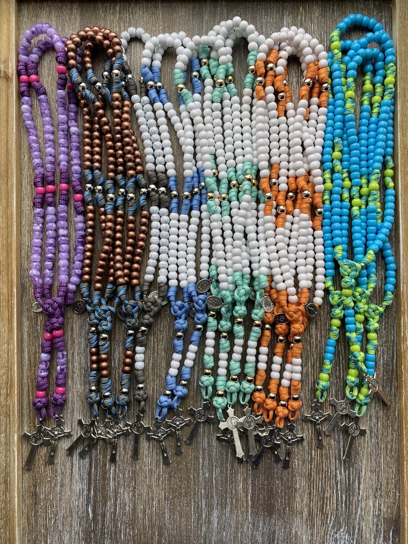 Puede incluir: Una colecci&oacute;n de siete rosarios con cuentas de pl&aacute;stico de colores, espaciadores met&aacute;licos y secciones de cord&oacute;n anudado. Cada rosario incluye un crucifijo de metal en la parte inferior y una peque&ntilde;a medalla circular, presentando combinaciones de colores como morado, bronce, azul, blanco, menta, naranja y azul brillante.