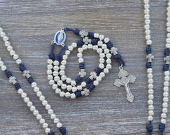May include: A rosary featuring silver-toned beads and dark blue cord knots. The design includes small silver cross spacers, a central oval medal with a blue inlay depicting a figure, and a detailed silver crucifix pendant at the end of the strand.