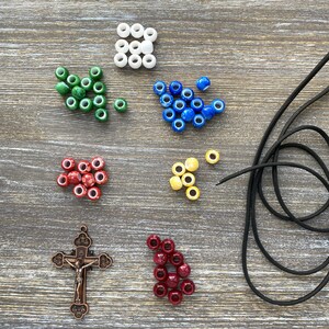 Puede incluir: Un conjunto de suministros para manualidades que incluye cuentas blancas, verdes, azules, amarillas, rojas y rojo oscuro, un colgante de crucifijo de metal en tono bronce con la figura de Jes&uacute;s y un cord&oacute;n negro fino para joyer&iacute;a.
