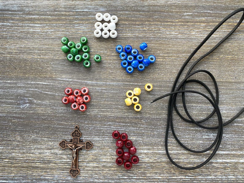 Puede incluir: Un conjunto de suministros para manualidades que incluye cuentas blancas, verdes, azules, amarillas, rojas y rojo oscuro, un colgante de crucifijo de metal en tono bronce con la figura de Jes&uacute;s y un cord&oacute;n negro fino para joyer&iacute;a.