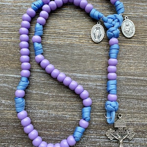 Puede incluir: Un rosario con cuentas lisas de color morado claro ensartadas en un cord&oacute;n azul brillante. El dise&ntilde;o incluye dos peque&ntilde;as medallas ovaladas de tono plateado con figuras religiosas y un crucifijo plateado, unidos por nudos de cord&oacute;n azul.