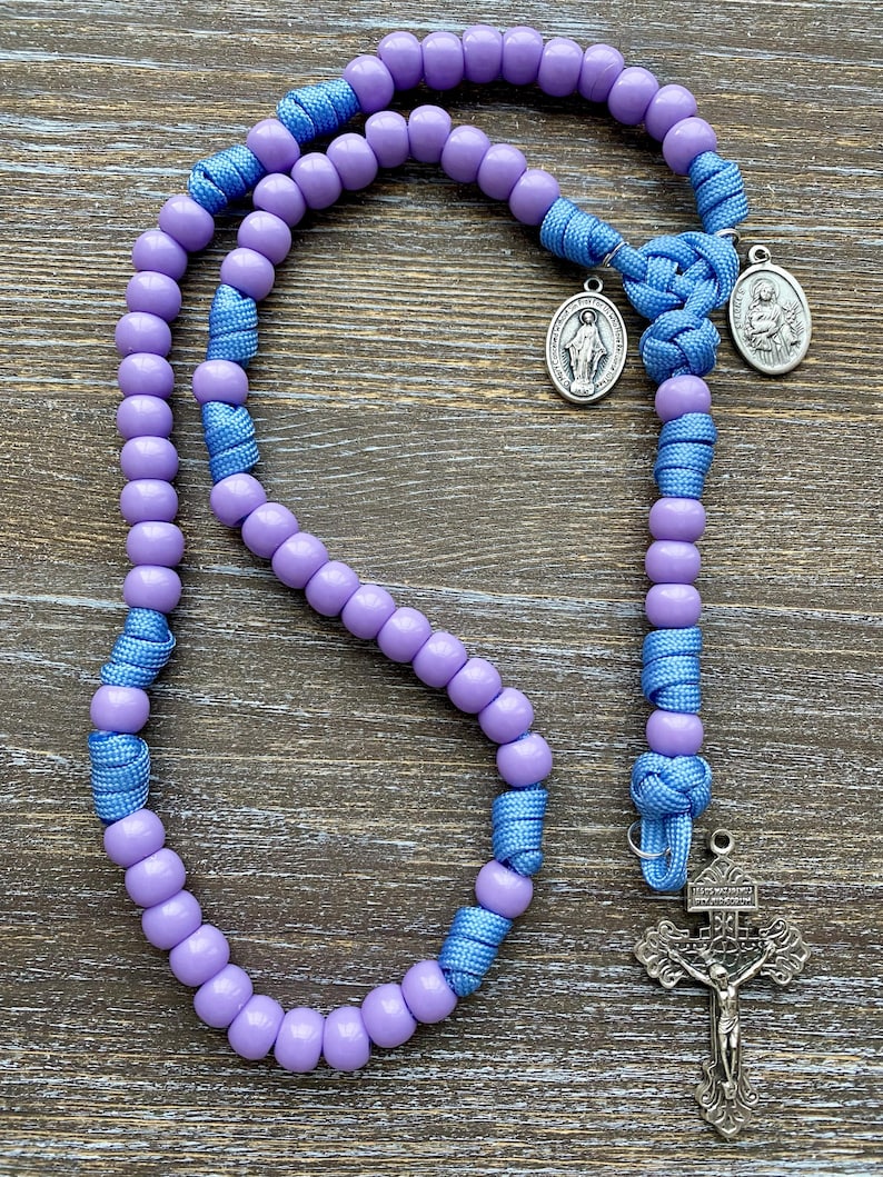 Puede incluir: Un rosario con cuentas lisas de color morado claro ensartadas en un cord&oacute;n azul brillante. El dise&ntilde;o incluye dos peque&ntilde;as medallas ovaladas de tono plateado con figuras religiosas y un crucifijo plateado, unidos por nudos de cord&oacute;n azul.