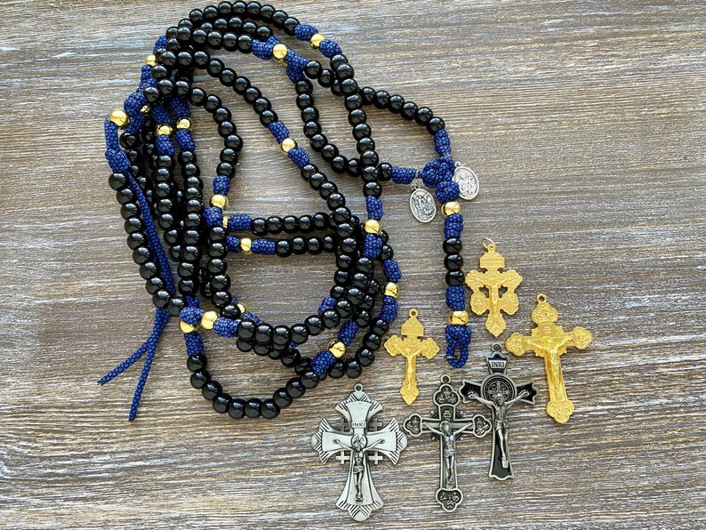 Puede incluir: Un rosario largo con cuentas negras lisas, detalles de cord&oacute;n azul y peque&ntilde;os espaciadores dorados. Dos medallas plateadas est&aacute;n unidas al rosario. Junto a &eacute;l hay cinco crucifijos separados con acabados met&aacute;licos en tonos oro y plata.