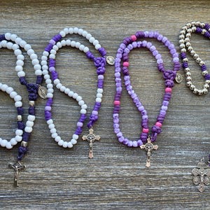 Puede incluir: Cuatro rosarios dispuestos en fila. El primero presenta cuentas blancas con detalles de cord&oacute;n morado y un crucifijo plateado. El segundo tiene cuentas blancas y moradas con un crucifijo plateado. El tercero usa cuentas en tonos morado y rosa con un crucifijo plateado. El cuarto tiene cuentas plateadas met&aacute;licas, cord&oacute;n morado y un crucifijo plateado.