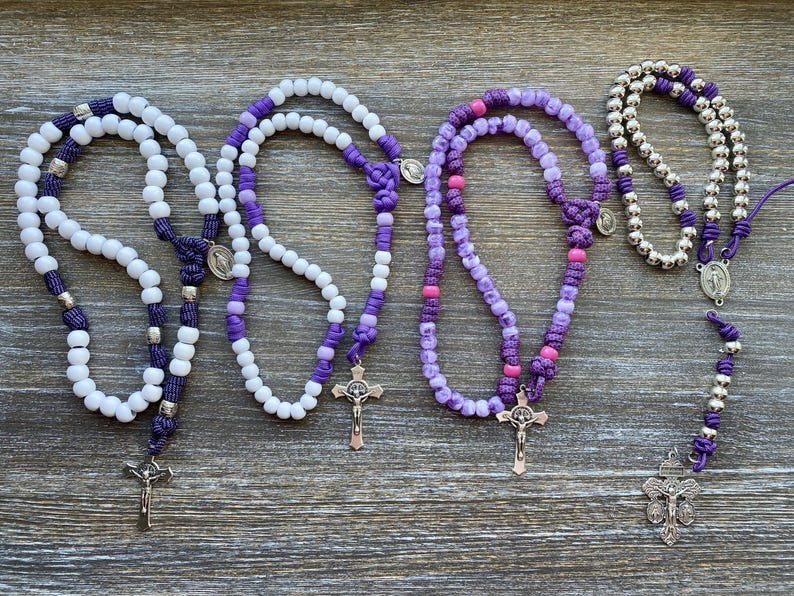 Puede incluir: Cuatro rosarios dispuestos en fila. El primero presenta cuentas blancas con detalles de cord&oacute;n morado y un crucifijo plateado. El segundo tiene cuentas blancas y moradas con un crucifijo plateado. El tercero usa cuentas en tonos morado y rosa con un crucifijo plateado. El cuarto tiene cuentas plateadas met&aacute;licas, cord&oacute;n morado y un crucifijo plateado.