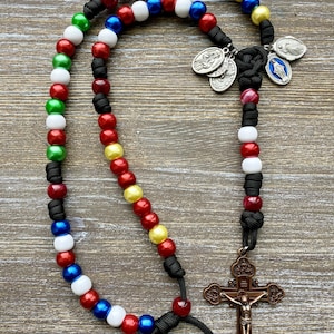 Puede incluir: Un rosario con cuentas multicolores en rojo, blanco, azul, verde y amarillo, ensartadas en un cord&oacute;n negro. El rosario incluye un crucifijo de tono bronce y varias medallas religiosas peque&ntilde;as de metal en tono plateado.