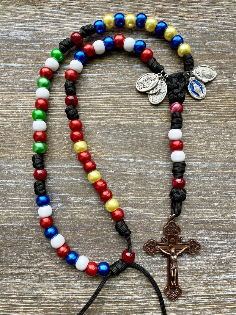 Puede incluir: Un rosario con cuentas multicolores en rojo, blanco, azul, verde y amarillo, ensartadas en un cord&oacute;n negro. El rosario incluye un crucifijo de tono bronce y varias medallas religiosas peque&ntilde;as de metal en tono plateado.