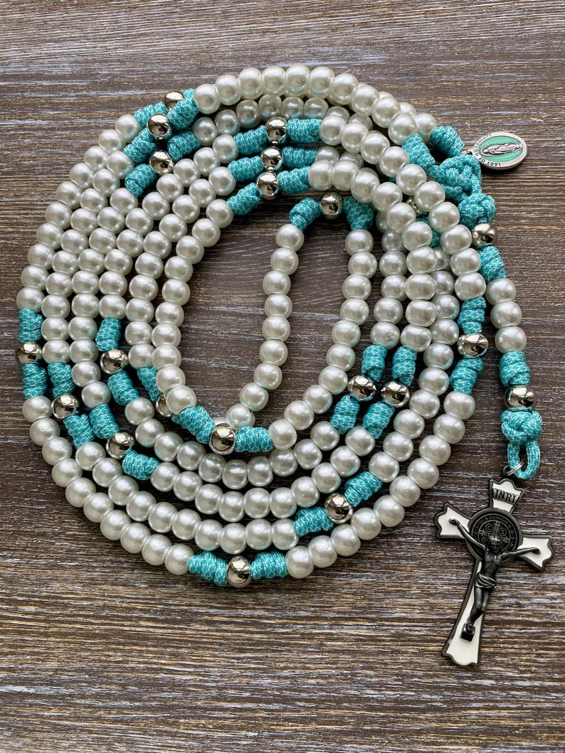 Puede incluir: Un rosario con cuentas de perlas blancas de imitaci&oacute;n y nudos de cord&oacute;n turquesa brillante, acentuado con cuentas de metal plateado pulido. El rosario incluye un crucifijo plateado con incrustaciones de esmalte blanco y una peque&ntilde;a medalla circular.