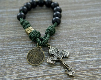 Puede incluir: Una pulsera de rosario con cuentas esféricas de color negro mate, cordón de paracaídas verde oliva con nudos intrincados, una cuenta metálica cuadrada con una cruz, una medalla redonda de San Benito y un crucifijo de bronce con la figura de Jesús.