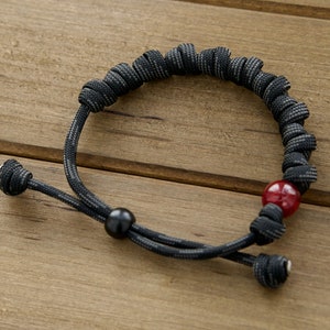 May include: A black and grey patterned paracord bracelet featuring a series of tight knots along the band. The bracelet includes one smooth red bead and one smooth black bead, with adjustable cord ends finished with small knots at the tips.