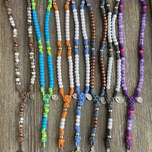 Puede incluir: Siete rosarios con cuentas de varios colores, incluyendo marr&oacute;n, azul, naranja, blanco y morado. Cada rosario est&aacute; construido con cord&oacute;n anudado, cuentas met&aacute;licas, una peque&ntilde;a medalla circular y un crucifijo de tono plateado.
