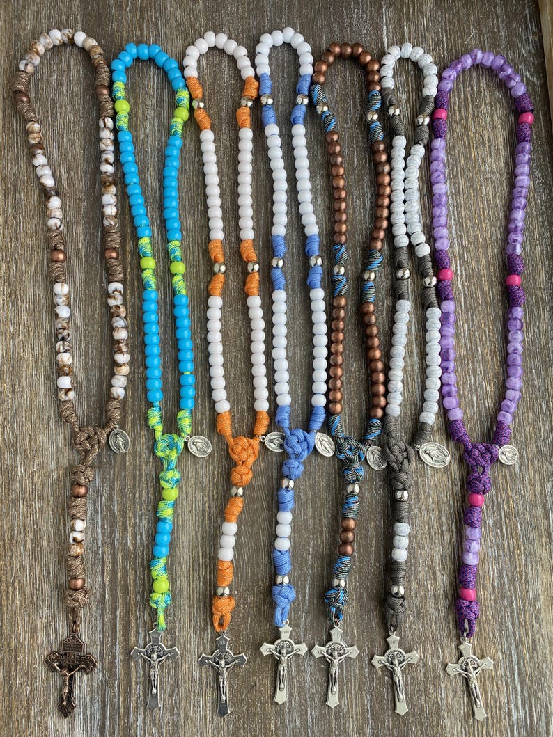 Puede incluir: Siete rosarios con cuentas de varios colores, incluyendo marr&oacute;n, azul, naranja, blanco y morado. Cada rosario est&aacute; construido con cord&oacute;n anudado, cuentas met&aacute;licas, una peque&ntilde;a medalla circular y un crucifijo de tono plateado.