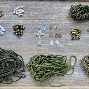 Puede incluir: Un conjunto de suministros para bisuter&iacute;a que incluye tres manojos de cord&oacute;n verde oliva, varias cuentas met&aacute;licas y blancas, un colgante de crucifijo plateado y varios dijes religiosos peque&ntilde;os en tonos plata y bronce.