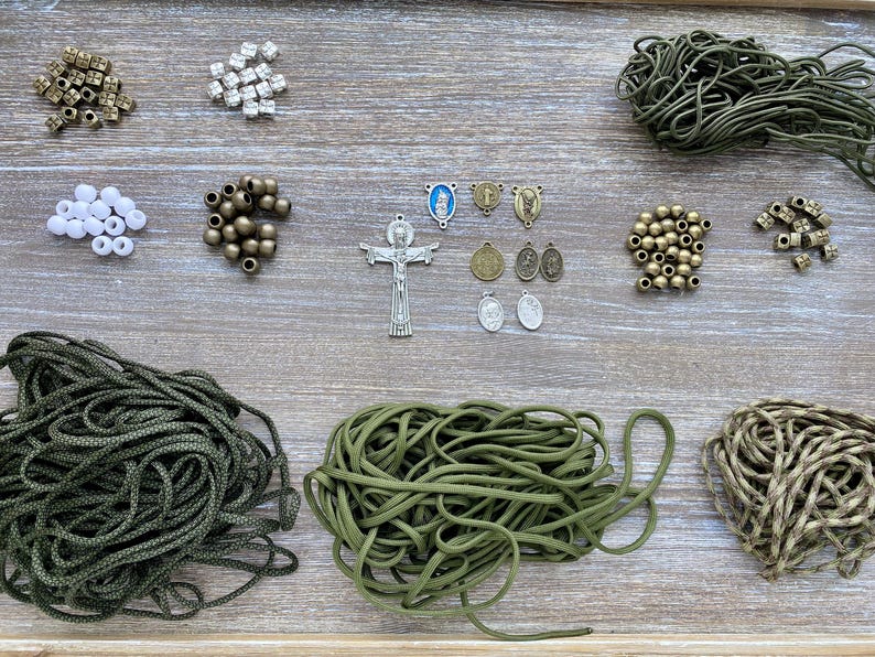 Puede incluir: Un conjunto de suministros para bisuter&iacute;a que incluye tres manojos de cord&oacute;n verde oliva, varias cuentas met&aacute;licas y blancas, un colgante de crucifijo plateado y varios dijes religiosos peque&ntilde;os en tonos plata y bronce.