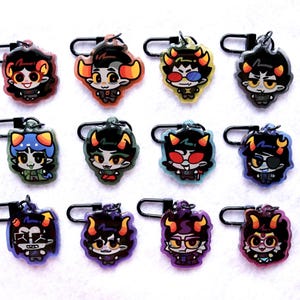 May include: A collection of twelve acrylic charms featuring stylised cartoon characters with various hair colours, horns, and expressions. Each charm is attached to a black metal lobster clasp, showcasing diverse designs including glasses, eye patches, and colourful outfits.