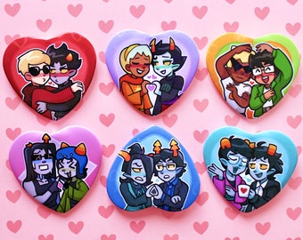 May include: A collection of six heart-shaped buttons featuring cartoon illustrations of couples. Each button has a different color scheme, with characters in various poses. The background is a light pink with small heart patterns.