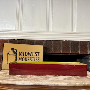 May include: A rectangular ceramic tray with a glossy deep red exterior and a bright lime green interior. Positioned behind the tray is a rectangular sign with a yellow background featuring a black jug icon and the text MIDWEST MODESTIES.