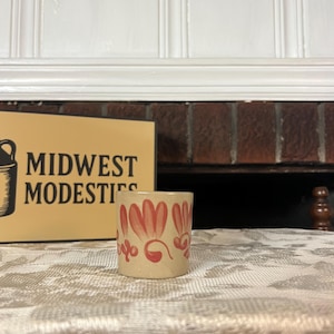 May include: A small, cylindrical beige ceramic cup featuring a hand-painted pattern of red, petal-like shapes and swirling flourishes around the exterior. The cup is positioned in front of a tan sign with the black text MIDWEST MODESTIES.