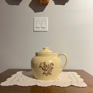 May include: A cream-colored ceramic teapot with a lid and handle, featuring a brown floral design. The teapot sits on a white lace doily on a wooden table. Woven baskets hang above the table against a gray wall.
