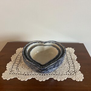May include: Set of three heart-shaped ceramic bowls with a speckled blue and white design. The bowls are nested on a cream-colored, crocheted doily. The bowls are on a dark brown wooden surface.