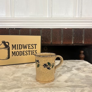 May include: A speckled beige ceramic mug featuring a hand-painted blue and green floral vine pattern around the center and a dark blue rim. The mug has a curved handle and a cylindrical body. Beside it sits a sign reading MIDWEST MODESTIES.
