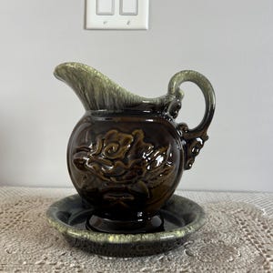 Könnte beinhalten: Ein Vintage-Set aus brauner Keramik, bestehend aus Krug und Schüssel. Der Krug hat ein Rosendesign und einen Griff mit dekorativen Elementen. Das Set hat eine glänzende Oberfläche mit einer zweifarbigen braun-grünen Glasur. Die Schüssel ist rund und farblich passend.