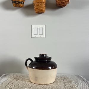 May include: A vintage stoneware jug with a dark brown top and handle, and a cream-coloured base. The jug has a lid and sits on a lace doily. Three small woven baskets hang above the jug.