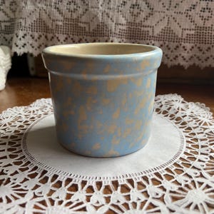 May include: A small, round, blue and tan speckled ceramic pot with a light tan interior. The pot sits on a white lace doily. The background includes a wooden surface and a white lace curtain.