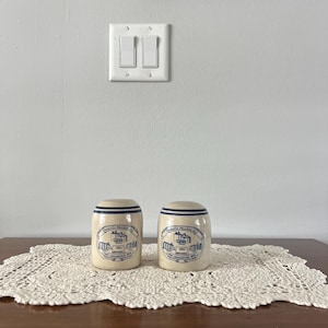 May include: Two ceramic salt and pepper shakers with a cream-colored base and blue accents. Each shaker has a blue band around the top and a printed design with text. They sit on a white lace doily on a wooden table.