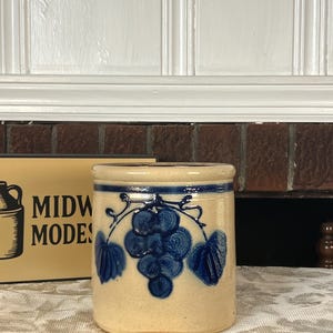 Pode incluir: Um pote de grés cilíndrico com acabamento bege e um design de cacho de uvas pintado à mão em azul cobalto. O recipiente apresenta uma borda espessa e uma faixa azul horizontal que circunda a parte superior, num estilo de sal clássico.