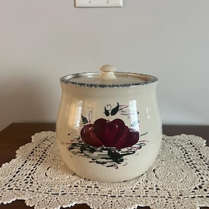May include: Cream-colored ceramic jar with a lid, featuring a hand-painted design of red apples with green leaves. The jar has a blue rim and sits on a lace doily. A decorative kitchen storage container.