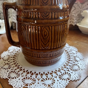 May include: A brown ceramic jug with a handle and spout, featuring a wood-grain design. The jug sits on a white lace doily. The jug is approximately 20 cm tall.