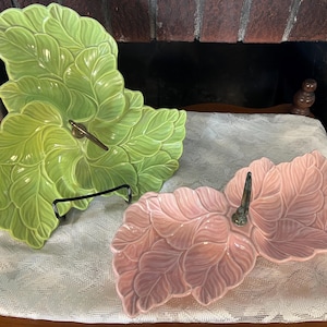 May include: Two ceramic serving dishes shaped like clusters of leaves. One dish is light green and features a central metal handle, while the other is light pink and also includes a central metal handle. Both pieces have a glossy finish.