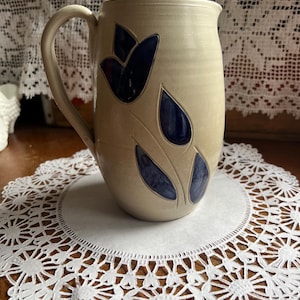 May include: A beige ceramic mug with a large handle, decorated with dark blue tulip-like designs. The mug sits on a white paper doily with a decorative lace edge. The mug is approximately 6 inches tall.