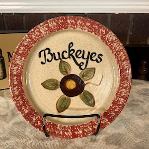 May include: A round ceramic plate with a speckled red and cream rim. The center features a hand-painted brown and gold buckeye nut surrounded by five green leaves. The word Buckeyes is written in black cursive script above the painted design.