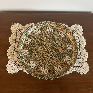 May include: A round, speckled green and tan decorative plate with white numbers marking increments of ten. The plate sits on a cream-colored, crocheted doily, placed on a dark wooden surface.