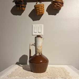 May include: A brown ceramic pitcher with a white and tan top, a handle, and vertical ridges. The pitcher sits on a white lace doily. Three small woven baskets hang on the wall above.