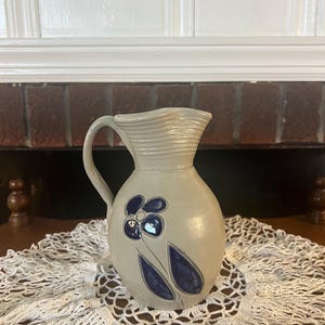 May include: A light beige ceramic pitcher featuring a ribbed neck and a single handle. The body of the pitcher is decorated with a dark blue, hand-painted floral motif consisting of a five-petaled flower and two elongated leaves on a smooth surface.