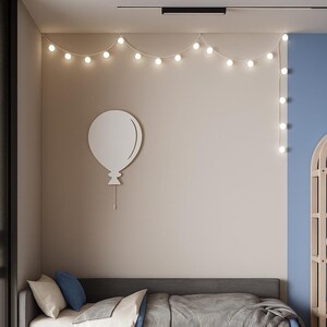 Op de afbeelding: Een moderne slaapkamer met een grijs bed, blauw en wit beddengoed en een grijze deken. Een witte ballon wanddecoratie en lichtslingers zijn zichtbaar. De kamer heeft een gestreept tapijt en houten vloer.